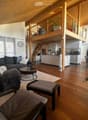 Open-concept living and dining area with wooden floors, a grey sectional sofa, coffee table, dining table, and kitchen area with white cabinets and appliances.