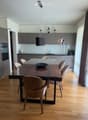 Modern kitchen with grey cabinets, a glass dining table, six chairs, a bowl on the table, and a hanging pendant light. The kitchen features a sleek design with a wooden floor and large windows with curtains.