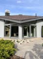 Modern single-story house with white exterior, large glass sliding doors, and a tiled roof. The house features a raised ground floor and is surrounded by neatly trimmed hedges and gravel pathways.
