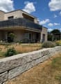 Modern two-story house with a balcony, patio seating area, and garden with stone wall and plants.