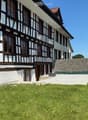 Traditional half-timbered house with white plaster walls, wooden beams, and multiple windows. The house has a sloped roof and is surrounded by a well-maintained lawn and a stone wall.