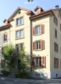 A multi-story residential house with light yellow walls, red shutters, and multiple windows. Features include a balcony on the second floor, a small garden area, and a staircase leading to the entrance.