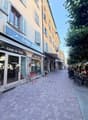 A charming pedestrian street lined with colorful buildings, featuring a clothing store named "Coups de Coeur" on the left and outdoor seating for a café on the right. The street is paved with cobblestones and has trees lining the far side.