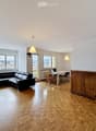 Open-plan living and dining area with wooden flooring, large windows offering a scenic view, a black leather sofa, a dining table with chairs, and a wooden cabinet.