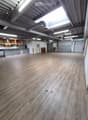Empty room with wooden flooring, white walls, and exposed industrial-style ceiling with pipes and lighting. Features include a hexagonal-patterned counter, a small seating area, and a doorway leading to another room.