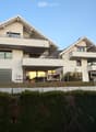 Modern two-story house with white exterior, large windows, balconies, and a garage. The house is situated on a raised ground floor with a well-maintained lawn and hedge in front.