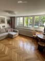 Living room with light wooden herringbone flooring, large windows with curtains, a cream sectional sofa with striped cushions, a wooden coffee table with a TV, and several framed artworks on the walls.