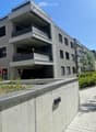 multistory building, balconies, gray walls, bricked pathway, shrubs, parking area in front