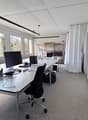 Modern office space with large windows, white walls, and a grey carpeted floor. A white desk holds a computer monitor, keyboard, mouse, and office supplies. An office chair is positioned in front of the desk. A black curtain is on the right side, and shel