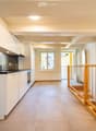 Modern hallway with tiled floor, white walls, built-in kitchen area on the left, staircase with wooden railing leading upwards, and a window at the end.
