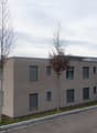 Modern two-story building with beige facade, multiple windows, and a balcony on the upper level. The building is surrounded by a few trees and a paved road.