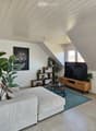 Modern living room with a white sofa, coffee table, TV stand, shelves, and potted plants. The room features a slanted wooden ceiling and large windows.