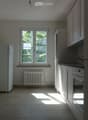 Empty kitchen with white cabinets, a window, a radiator, and a refrigerator. No furniture or appliances are present.