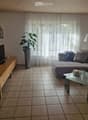 Modern living room with a grey sectional sofa, glass coffee table, flat-screen TV on a wooden cabinet, and a large plant in a grey vase. The room features tiled flooring and large windows with sheer curtains.