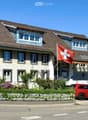 Two-story house with a brown tiled roof, white siding, multiple windows, and a Swiss flag. A red SUV is parked in front, and there is a small garden with a white picket fence.
