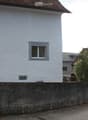 White plastered wall, brown roof, grey concrete fence, two windows, a window on the corner