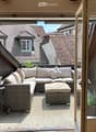 A modern balcony with a grey wicker sofa, ottoman, small table, and potted plants, overlooking a cityscape with traditional rooftops.
