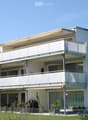 Modern white apartment building with multiple balconies and large windows, surrounded by a small garden area with potted plants.