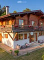 A two-story wooden house with a large balcony, surrounded by greenery and a well-maintained lawn. The house features a mix of wooden and white exterior walls, multiple windows with shutters, and a chimney. Outdoor seating includes a sofa, chairs, and a sm