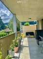 Balcony with a sofa, small table, chairs, potted plants, grill, and storage bin, overlooking a green landscape.