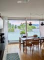 Open-plan kitchen and dining area with modern white cabinets, a black countertop, and a dining table with six chairs. The space features large windows with a view of greenery and a balcony area.