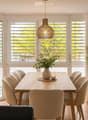 Bright dining area with a wooden table, six chairs, a vase with greenery, and a candle on the table. Large windows with blinds offer natural light and a view of greenery outside.