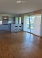 Empty room with wooden flooring, white walls, and large windows with blinds. Kitchen area visible in the background with cabinets and a countertop.
