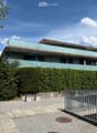 Modern two-story building with large glass windows and balconies, surrounded by greenery and a gravel area.