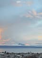 Panoramic cityscape view at sunset, featuring a body of water, distant mountains, and urban buildings.