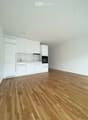 Empty room with light-colored walls, wooden flooring, and a small white kitchen area featuring cabinets and a sink.