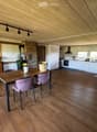 Open-plan living and dining area with wooden floors, exposed wooden beams, a dining table with chairs, a cabinet, and a modern kitchen area with white cabinets and stainless steel appliances.
