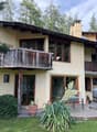 Two-story house with a wooden balcony, large windows, stone chimney, and outdoor seating area with a table and chairs.