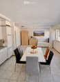 Bright, modern open-plan living and dining area with a fully equipped kitchen, dining table with chairs, TV stand, and a separate bedroom area with a bed and wardrobe.