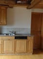 Compact kitchen with light wooden cabinets, granite countertop, sink, dishwasher, and refrigerator. Wooden floor and exposed wooden beams on the ceiling.