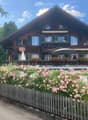 A two-story wooden house with green shutters, a balcony adorned with flowers, and a garden with pink roses and a wooden fence.