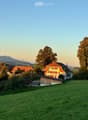 A cluster of traditional houses with red-tiled roofs and white walls, surrounded by lush green fields and tall trees, with distant mountains visible in the background.