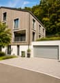 Modern two-story house with wooden siding, large windows, and balconies. Features a garage, paved driveway, and landscaped front yard with potted plants and a green waste bin.