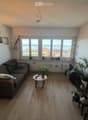Modern living room with large windows offering a scenic view, featuring a brown sectional sofa, a small wooden coffee table, a floor lamp, a tall shelving unit with books and plants, and decorative picture frames on the wall.