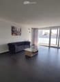 Modern living room with dark tiled flooring, grey sofa, glass coffee table, TV stand, and large sliding glass doors leading to a balcony.