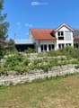 Two-story modern house with red-tiled roof, large windows, and a raised ground floor surrounded by a stone retaining wall and greenery.