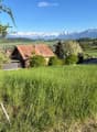 A scenic view of a grassy field with two houses in the background, surrounded by lush greenery and distant snow-capped mountains.