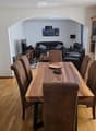 Dining area with a wooden dining table and chairs, a living area with a sofa and armchair, and a television. The room features wooden flooring and white walls.