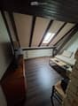 An attic room with wooden floors, exposed wooden beams, and a skylight. It features a sofa, coffee table, a wicker chair, a screen, and a small table with items on it.