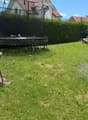Lush green garden with a trampoline, a wicker chair with cushions, and a large umbrella on a patio.