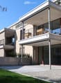Modern multi-story building with balconies, large windows, and a clean, contemporary design. Features include a raised ground floor, a well-maintained lawn, and a paved pathway leading to the entrance.