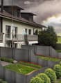 Two-story house with a tiled roof, multiple windows, and balconies. The surrounding area includes a well-maintained garden with a grey wooden fence, shrubs, and flower beds.