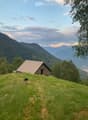 A small stone house with a metal roof sits on a grassy hillside, surrounded by trees and mountains.