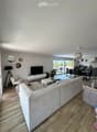 Bright, modern living room with large windows and a balcony. Features a grey sectional sofa, coffee table, armchair, and a dining area with a table and chairs.