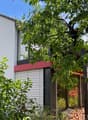 Modern two-story house with white walls, red accents, large windows, and a balcony. Surrounded by lush greenery and a stack of firewood near the entrance.