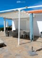 Outdoor patio with stone tiles, white pergola, sofa, table, chairs, and umbrella, overlooking the ocean.